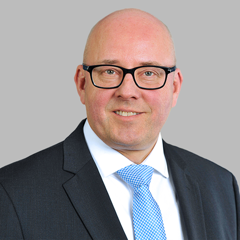 A bald man in a dark charcoal suit, white shirt and light blue patterned tie stands before a bright windowed office background.