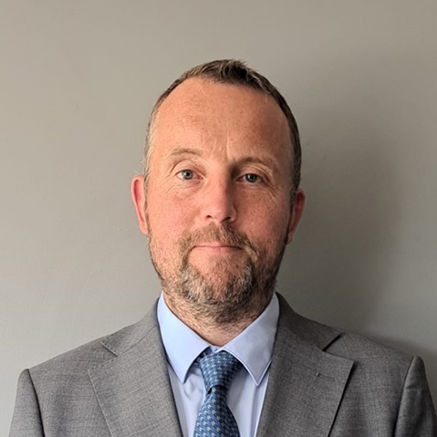 A person in a grey suit and blue tie stands against a plain background.