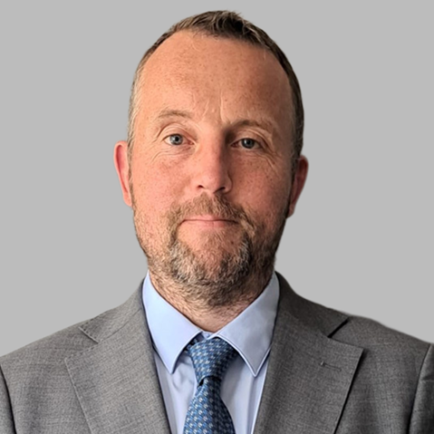 A person in a grey suit and blue tie stands against a plain background.